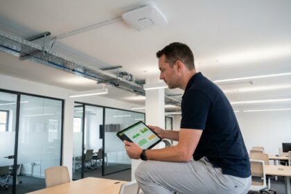 Technicien informatique inspectant une borne Wi-Fi 6E montée au plafond dans un bureau d'entreprise moderne avec une tablette affichant un tableau de bord réseau